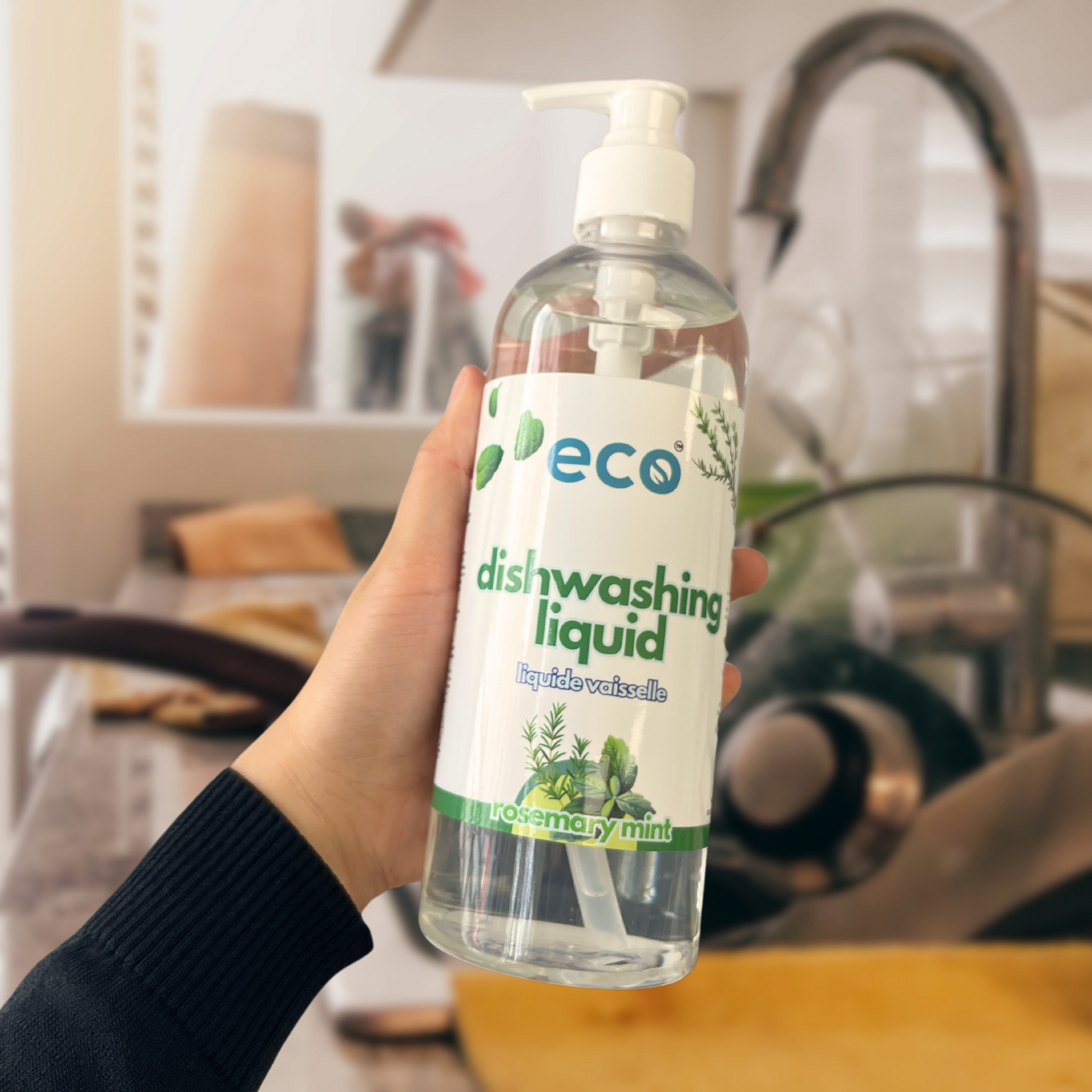 A bottle of eco dishwashing soap held in front of a sink full of dishes, for an eco-friendly and sustainable kitchen.